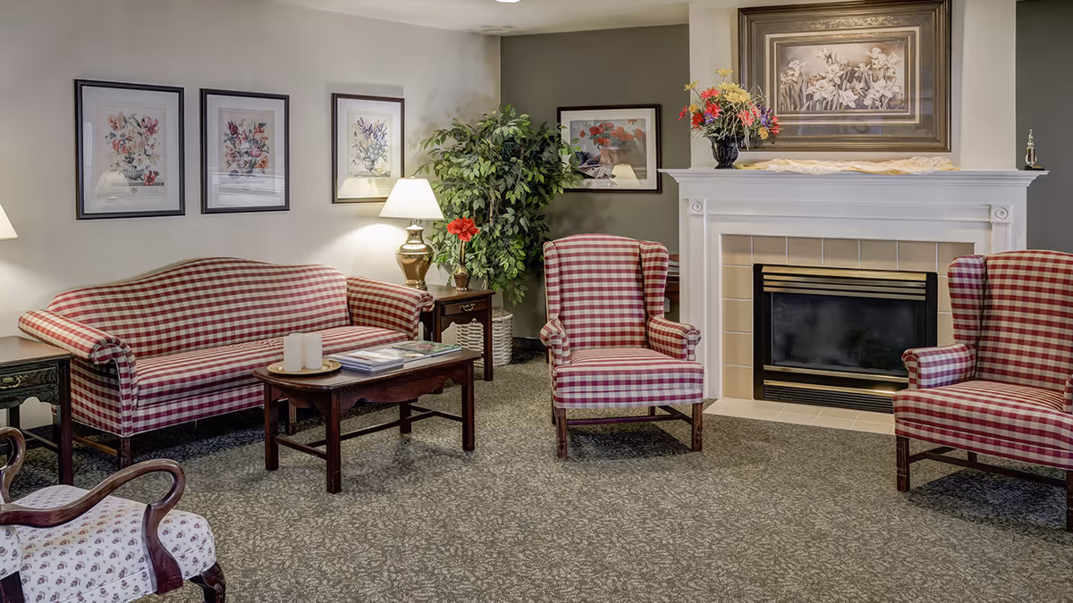 A cozy living room with a red and white checkered sofa and two matching armchairs arranged around a wooden coffee table. The room features a white fireplace with a floral painting above it and a vase of flowers on the mantel. There are framed floral artworks on the walls, a table lamp, and a large green plant in the corner.
