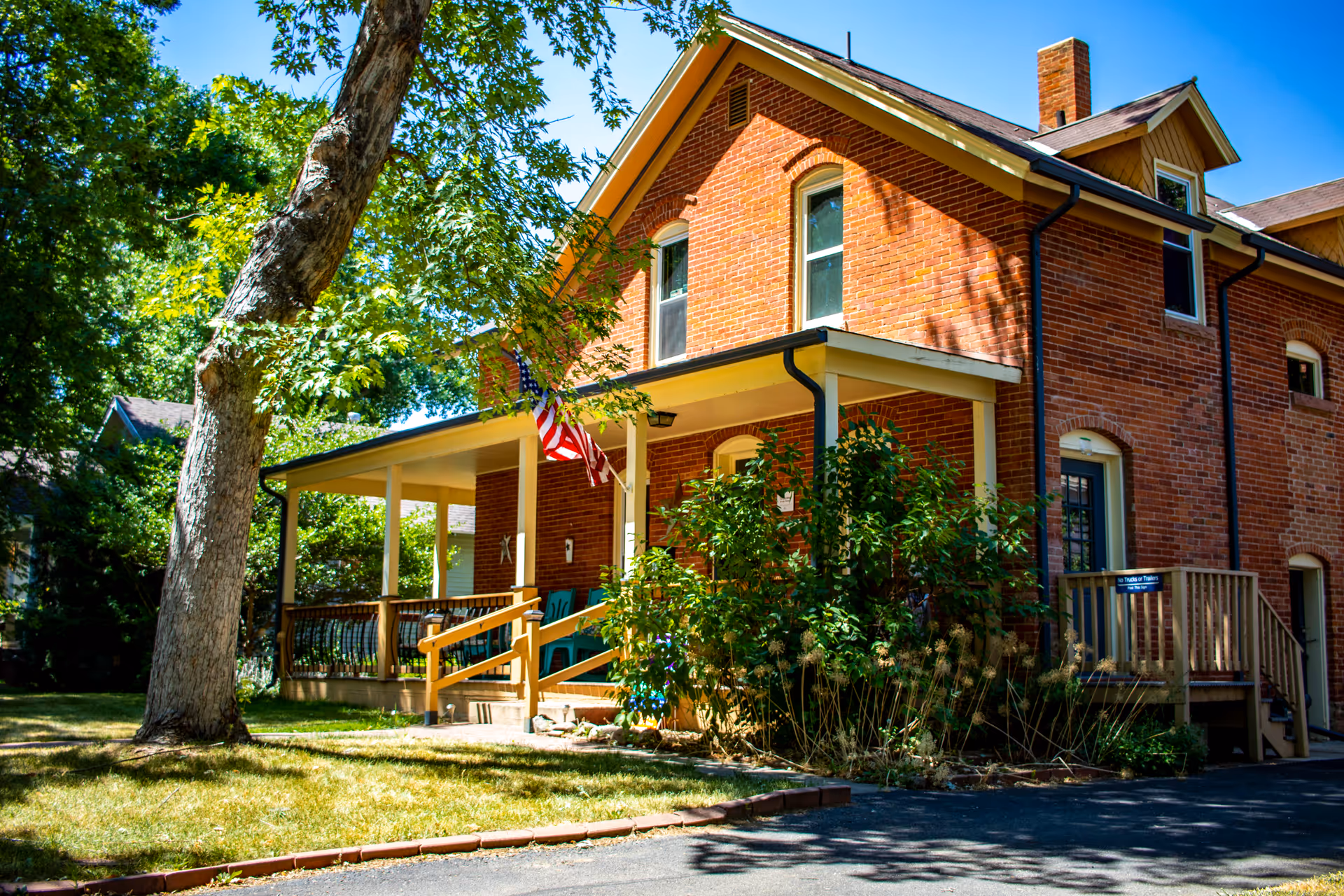 Two-story red brick house with a covered front porch, an American flag, and trees in the yard.