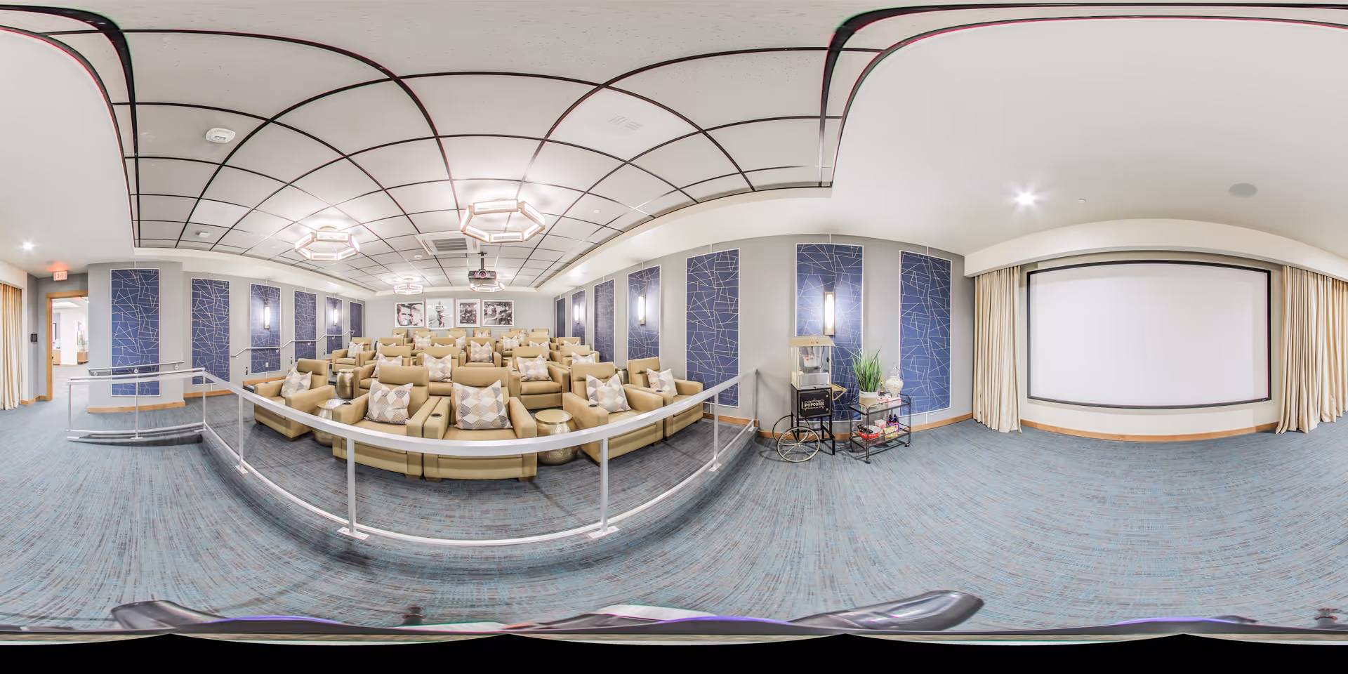 A spacious, modern theater room with multiple rows of beige recliner chairs, each with patterned cushions. The room features a large projection screen at the front, blue decorative wall panels, and soft lighting fixtures on the ceiling and walls. There is a popcorn machine and a small cart with plants and snacks near the screen. The room has a blue carpet and a white ceiling with a grid pattern.