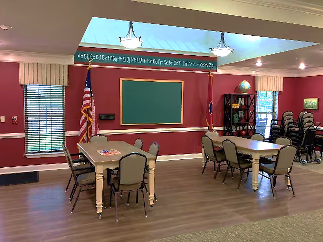 Interior room with red walls and two tables surrounded by chairs. There is a green chalkboard on the wall, an American flag, a Tennessee state flag, and a bookshelf with a globe and decorative letters. Windows with blinds and stacked chairs are visible in the background.