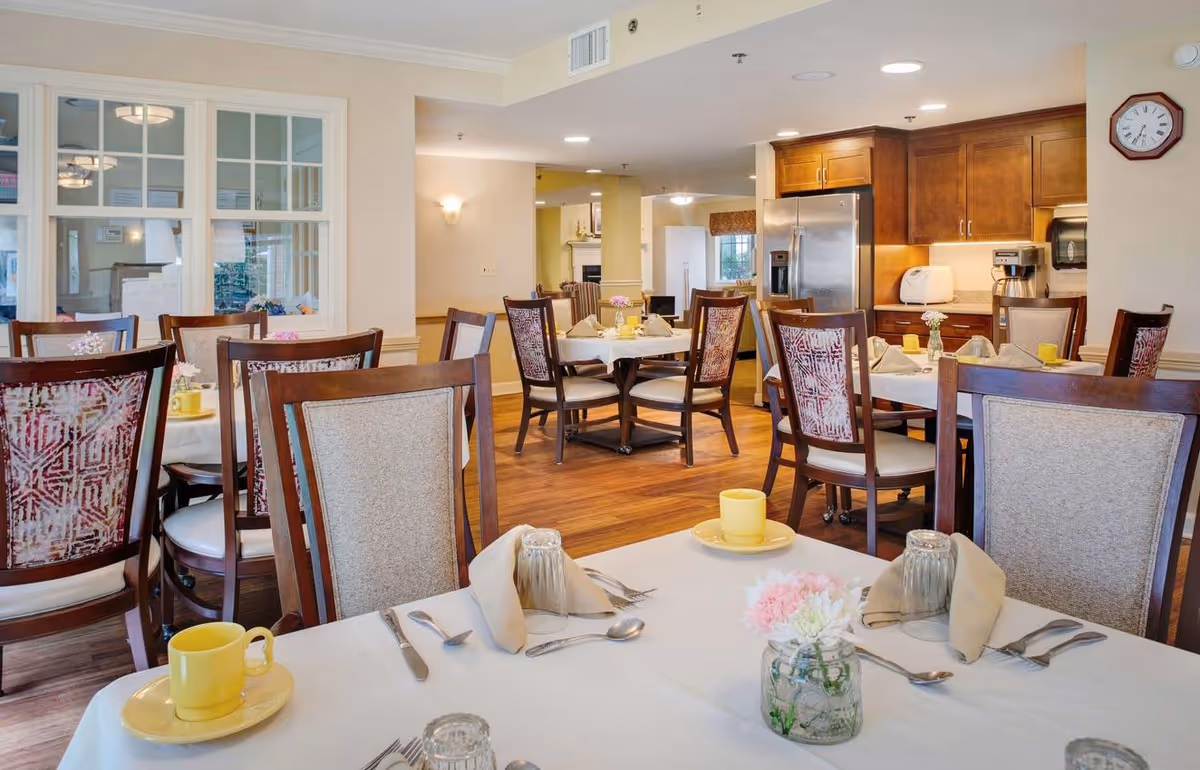 A bright communal dining room with multiple set tables and chairs and a kitchen area in the background.