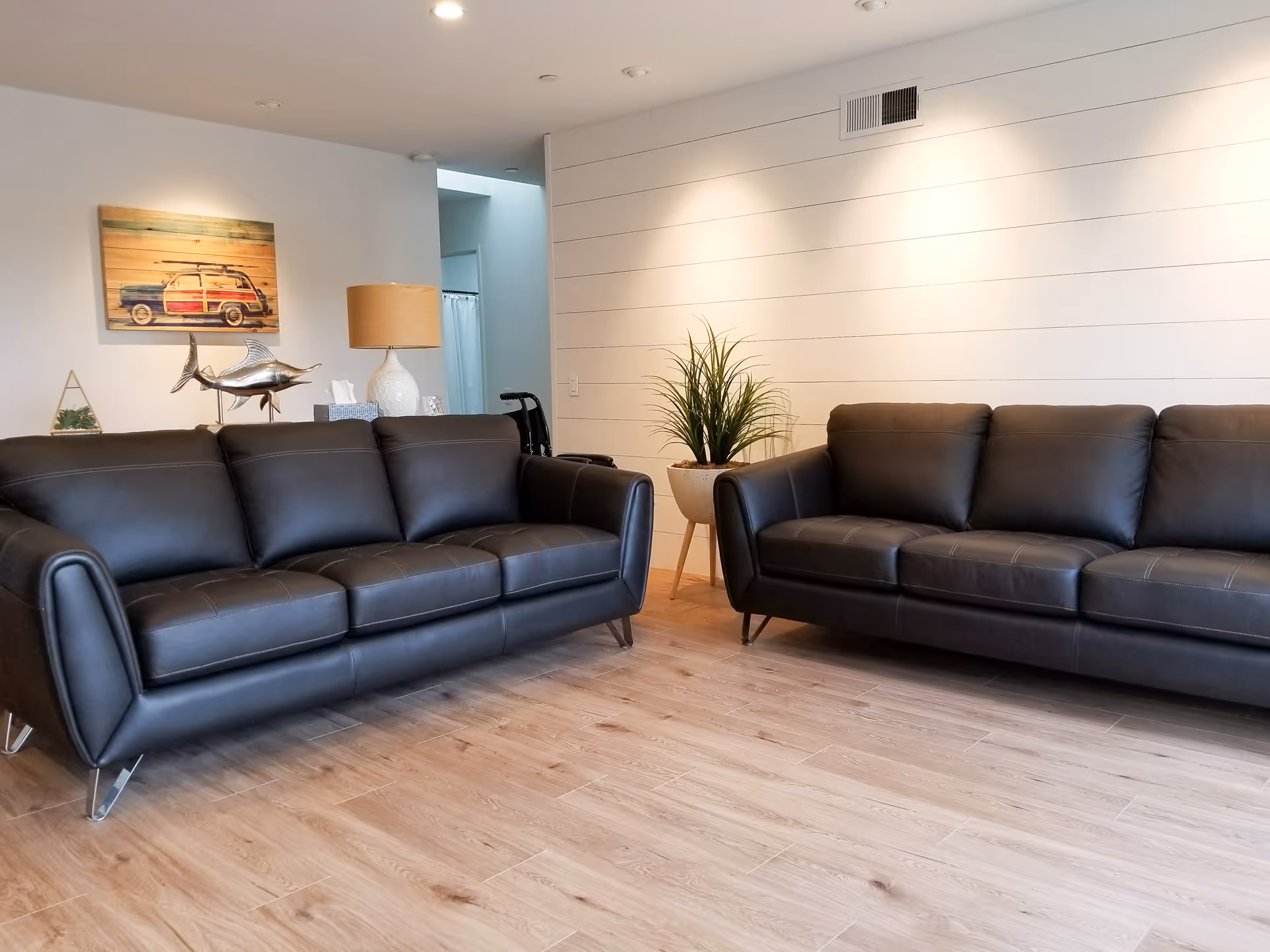 A modern living room with two black leather sofas facing each other on a light wooden floor. The walls are white with horizontal paneling, and there is a potted plant on a wooden stand between the sofas. A table lamp, a decorative fish sculpture, and a wall art piece featuring a vintage car are visible in the background.