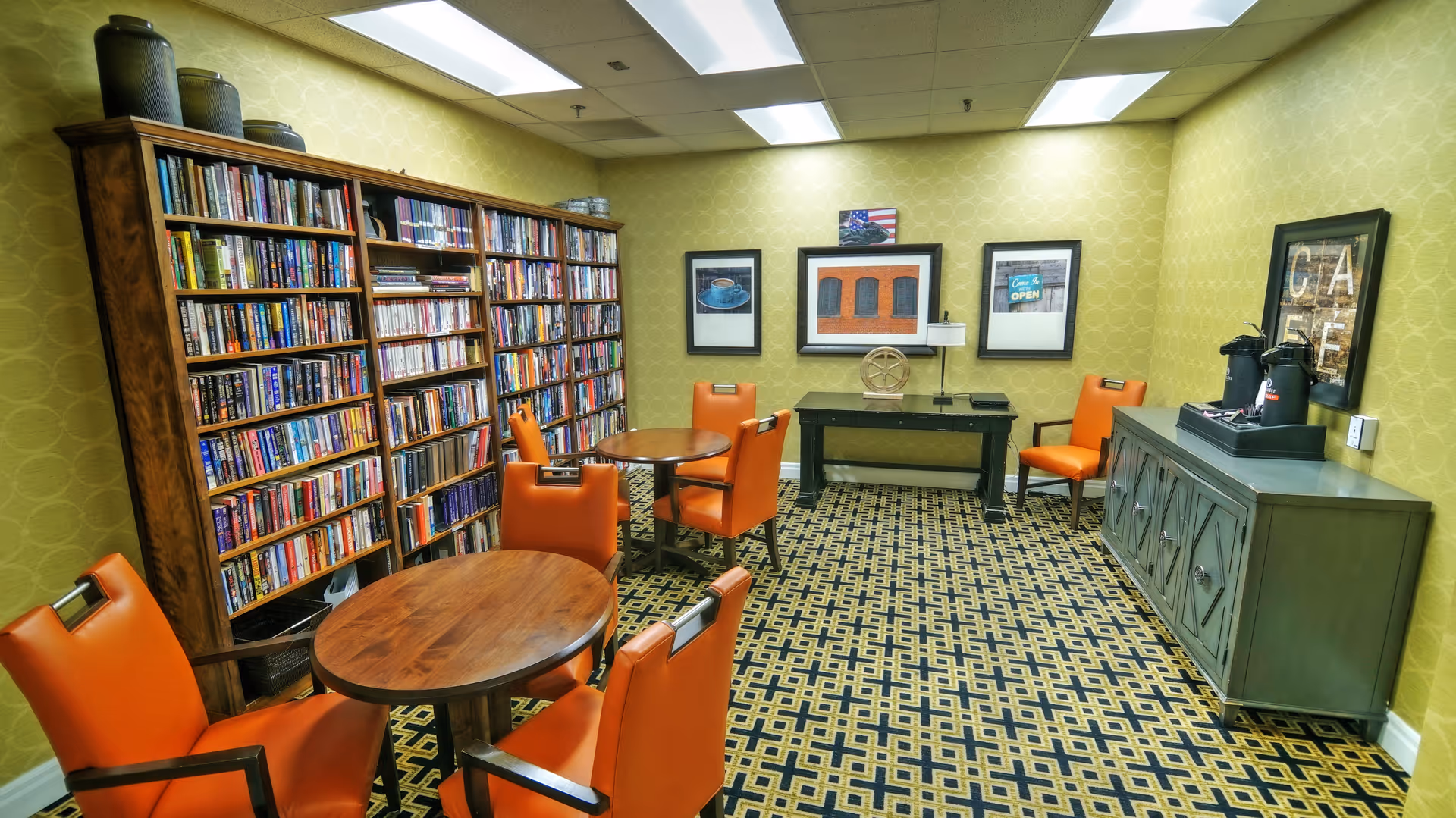 A cozy reading room with yellow patterned walls and a geometric carpet. The room features two round wooden tables surrounded by orange cushioned chairs. Along one wall is a large wooden bookshelf filled with books. On the opposite wall, there is a green sideboard with coffee dispensers and a framed 'CAFE' sign above it. Three framed pictures hang on the far wall above a small black table with decorative items.