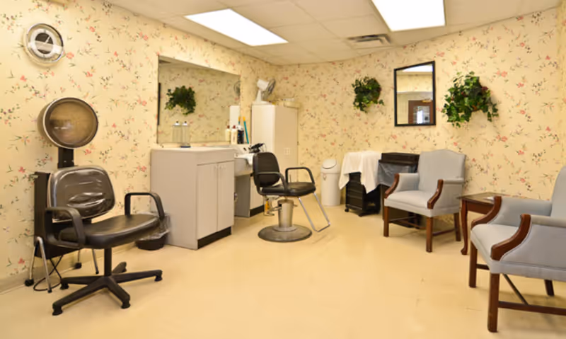 A small salon or beauty care room with two black salon chairs, a white cabinet with a sink, a large mirror, and floral wallpaper. There are two gray upholstered chairs with wooden armrests, a small wooden side table, and two wall-mounted green plants. The floor is light-colored and the ceiling has fluorescent lighting.