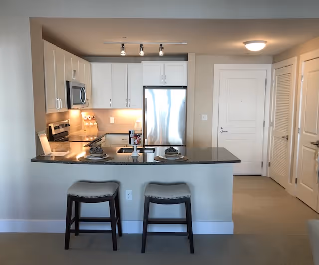Open-plan kitchen with a granite island, two stools, stainless steel refrigerator and white cabinets facing the entry door.