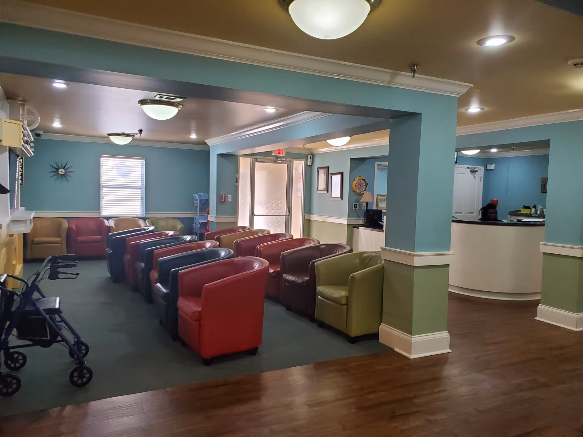 Interior view of a senior living facility waiting area with multiple colorful armchairs arranged in rows, a walker near the entrance, teal and green walls, and a reception desk with a staff member behind it.