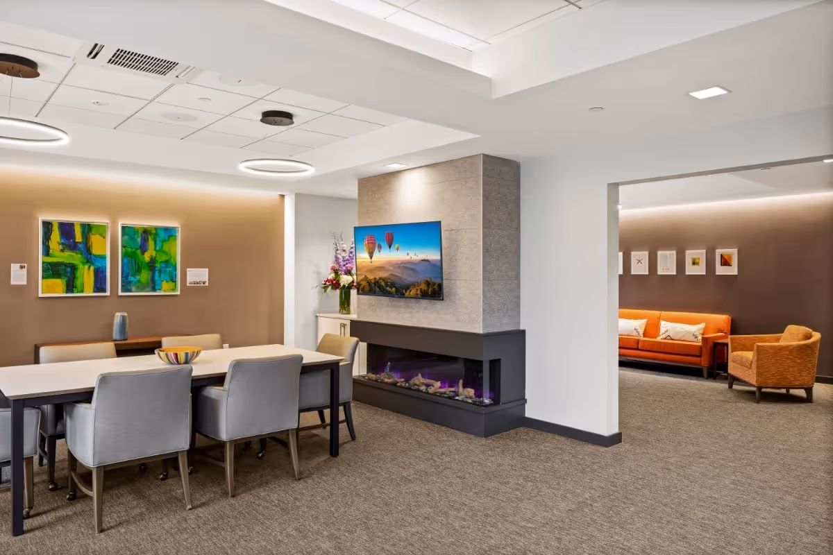 A modern interior space featuring a rectangular dining table with six gray upholstered chairs. On the table is a colorful bowl. Behind the table, there are two abstract paintings on a beige wall and a small sideboard with a vase. To the right, a contemporary fireplace with a mounted TV above it displays an image of hot air balloons over a scenic landscape. In the background, through an open doorway, there is a seating area with an orange sofa and an orange armchair against a brown wall with framed artwork.