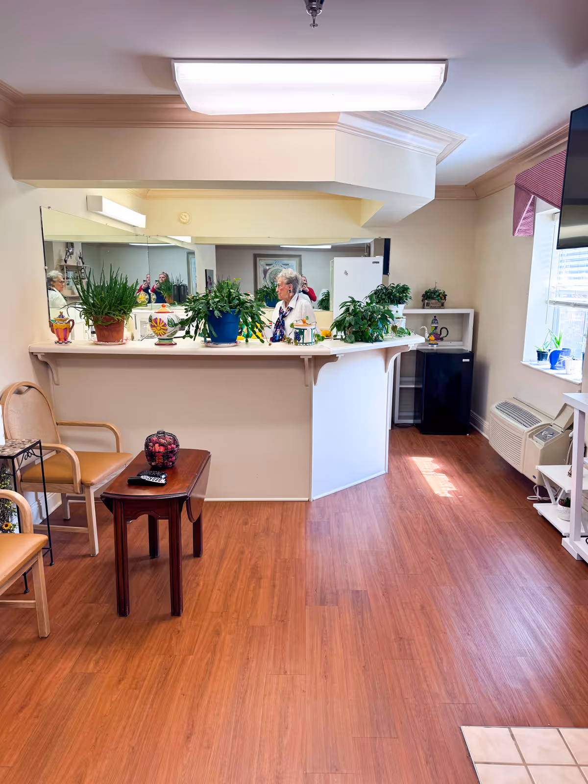 Interior view of a common area in Spring Terrace Assisted Living featuring a wooden floor, a white counter with several potted plants on top, a large mirror behind the counter, two chairs, a small wooden table with a decorative bowl and a remote control, a window with blinds and a mounted TV on the right wall, and an elderly person standing behind the counter.