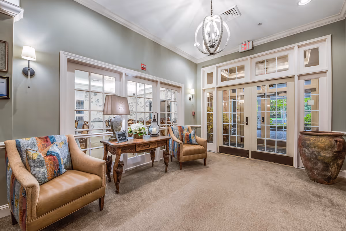 Well-lit lobby seating area with two upholstered chairs, a decorative table with a lamp and flowers, and glass double doors leading outside.