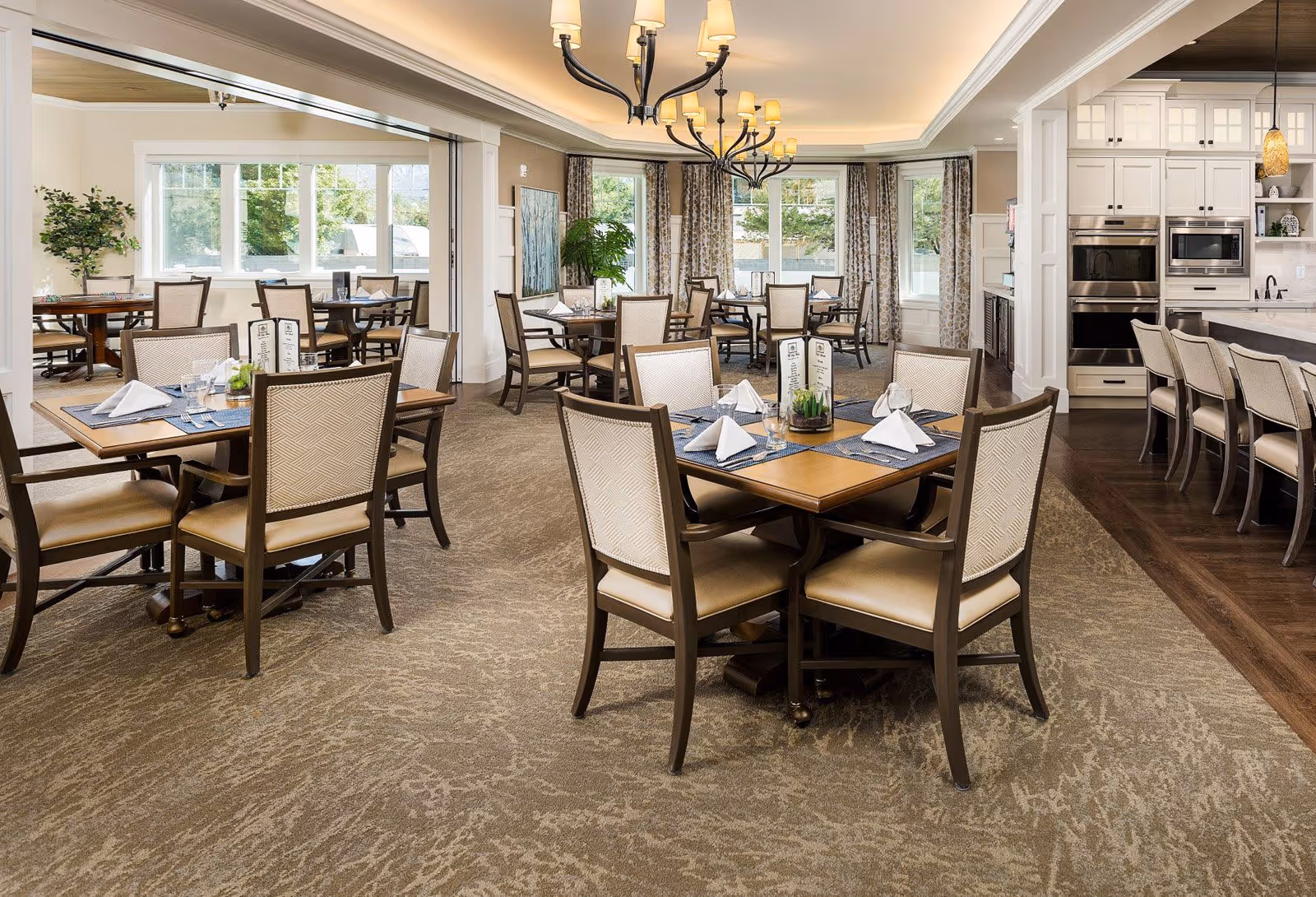 A bright and elegant dining room in a senior living facility with multiple wooden tables and cushioned chairs arranged neatly. Each table is set with folded white napkins, glasses, and menus. Large windows allow natural light to fill the room, and there are decorative plants and curtains along the walls. The room also features a kitchen area with white cabinetry and bar stools.
