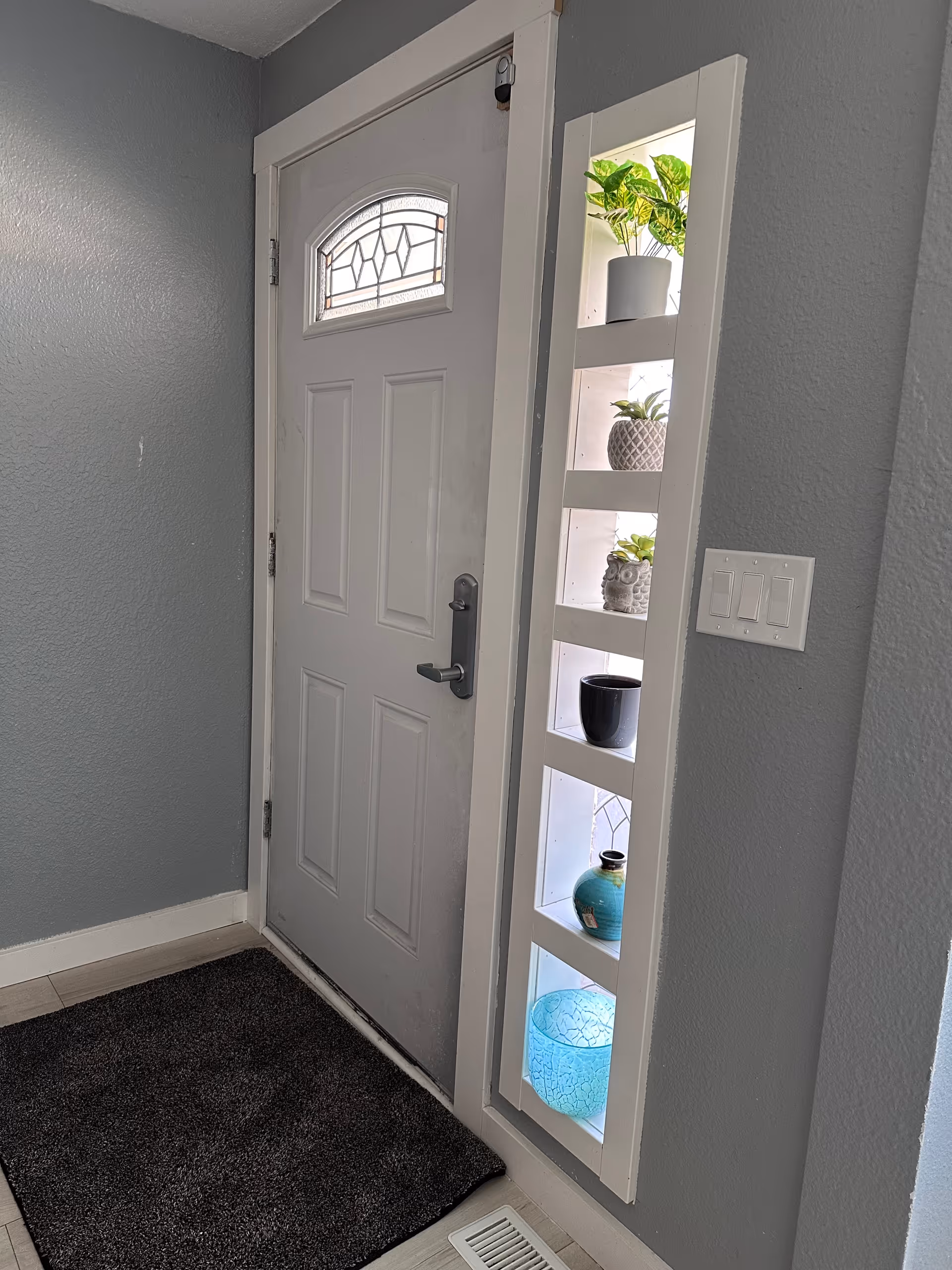 Interior view of an entryway featuring a closed white door with a decorative glass window at the top. To the right of the door is a built-in vertical shelf with five compartments, each holding decorative items including potted plants and vases. The walls are painted gray, and there is a dark gray doormat on the light-colored floor in front of the door. A light switch panel is visible on the wall next to the shelf.