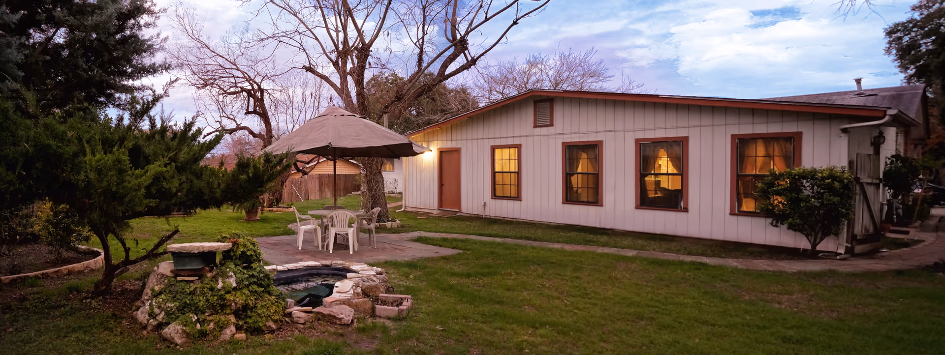Outdoor view of a single-story building with white siding and brown trim, featuring four windows with warm interior lighting. In the foreground, there is a small stone water feature surrounded by greenery, a patio area with a table, chairs, and a large umbrella. Trees and bushes are visible around the yard under a partly cloudy sky at dusk.