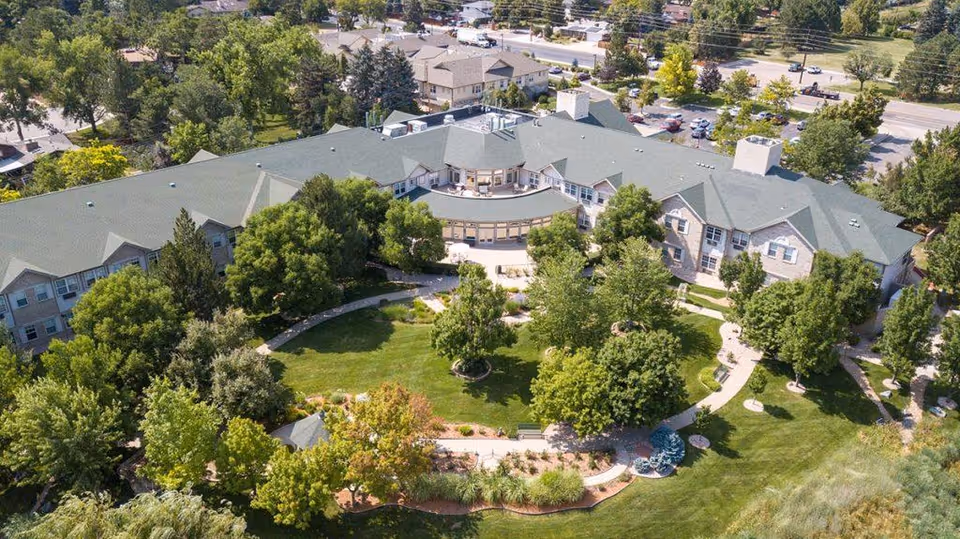 Aerial view of Gardens at Columbine senior living facility showing a large building surrounded by lush green trees, landscaped gardens, and walking paths. The building has a gray roof and multiple windows, with a curved patio area in the center. The surrounding area includes parking lots and additional greenery.