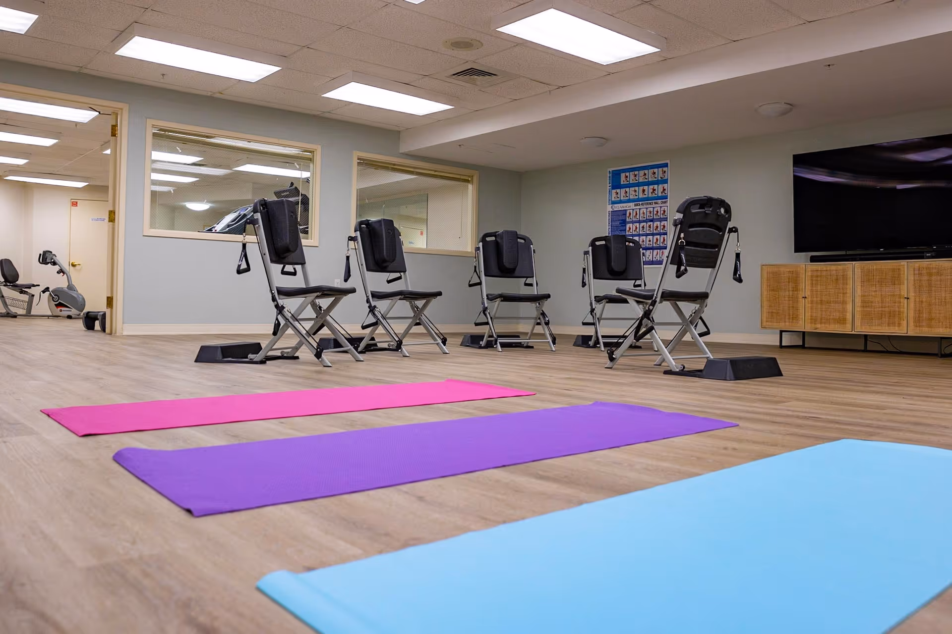A fitness/activity room with colorful yoga mats on the floor, foldable exercise chairs, and a TV cabinet along the wall.