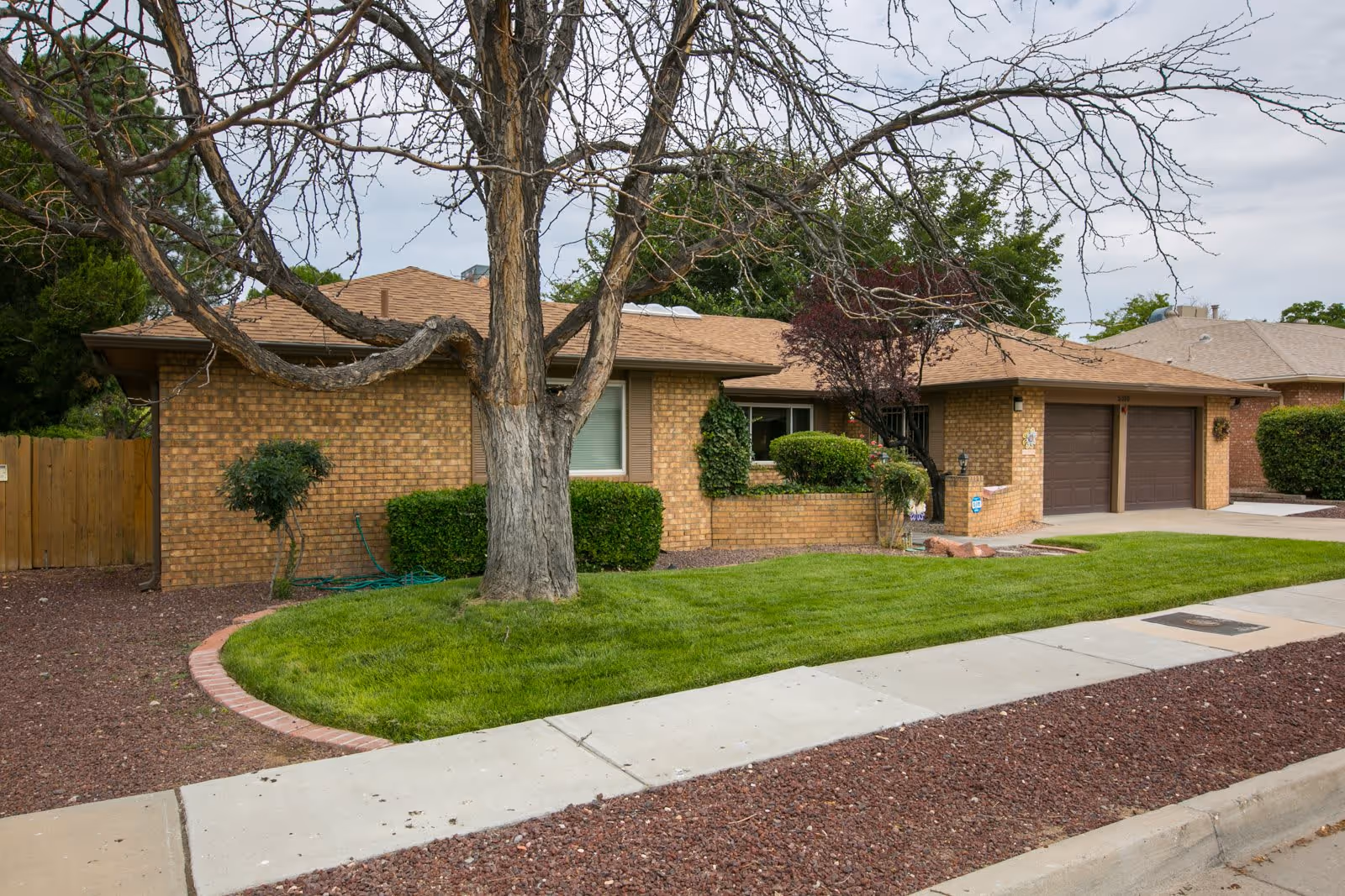 Exterior view of a single-story brick house with a brown shingled roof, a large tree with bare branches in the front yard, green grass, and a concrete sidewalk in front. The house has two brown garage doors and some bushes near the windows.