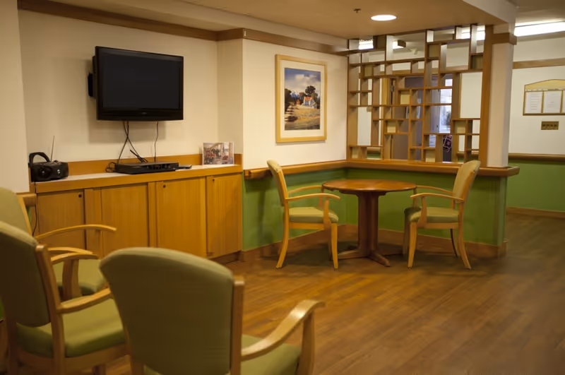 Communal sitting area with green-upholstered chairs around a round wooden table, a wall-mounted TV, and wood cabinetry.
