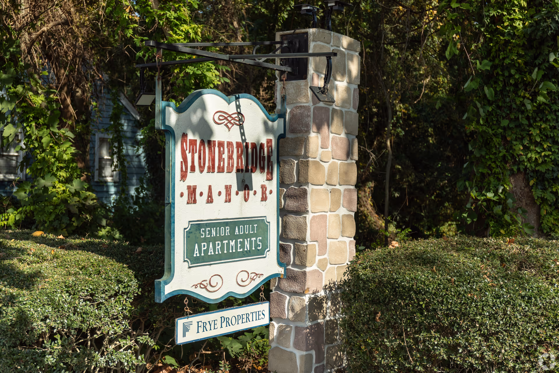 Hanging entrance sign reading "Stonebridge Manor Senior Adult Apartments" mounted on a stone pillar surrounded by shrubs and trees.