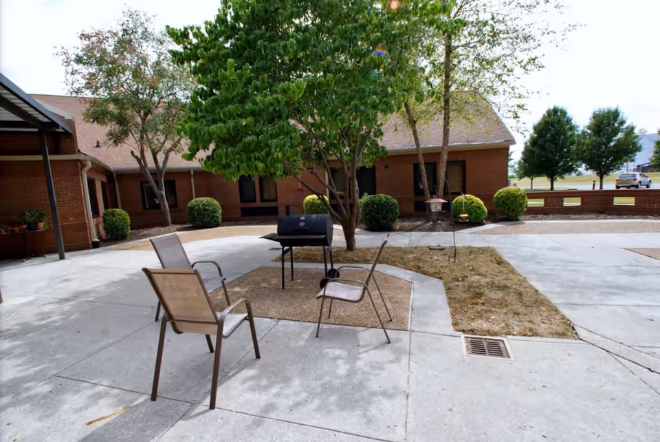 Outdoor courtyard area with a few metal chairs arranged around a black grill. There are trees and bushes planted in the middle and around the edges of the concrete patio. A brick building with windows surrounds the courtyard.