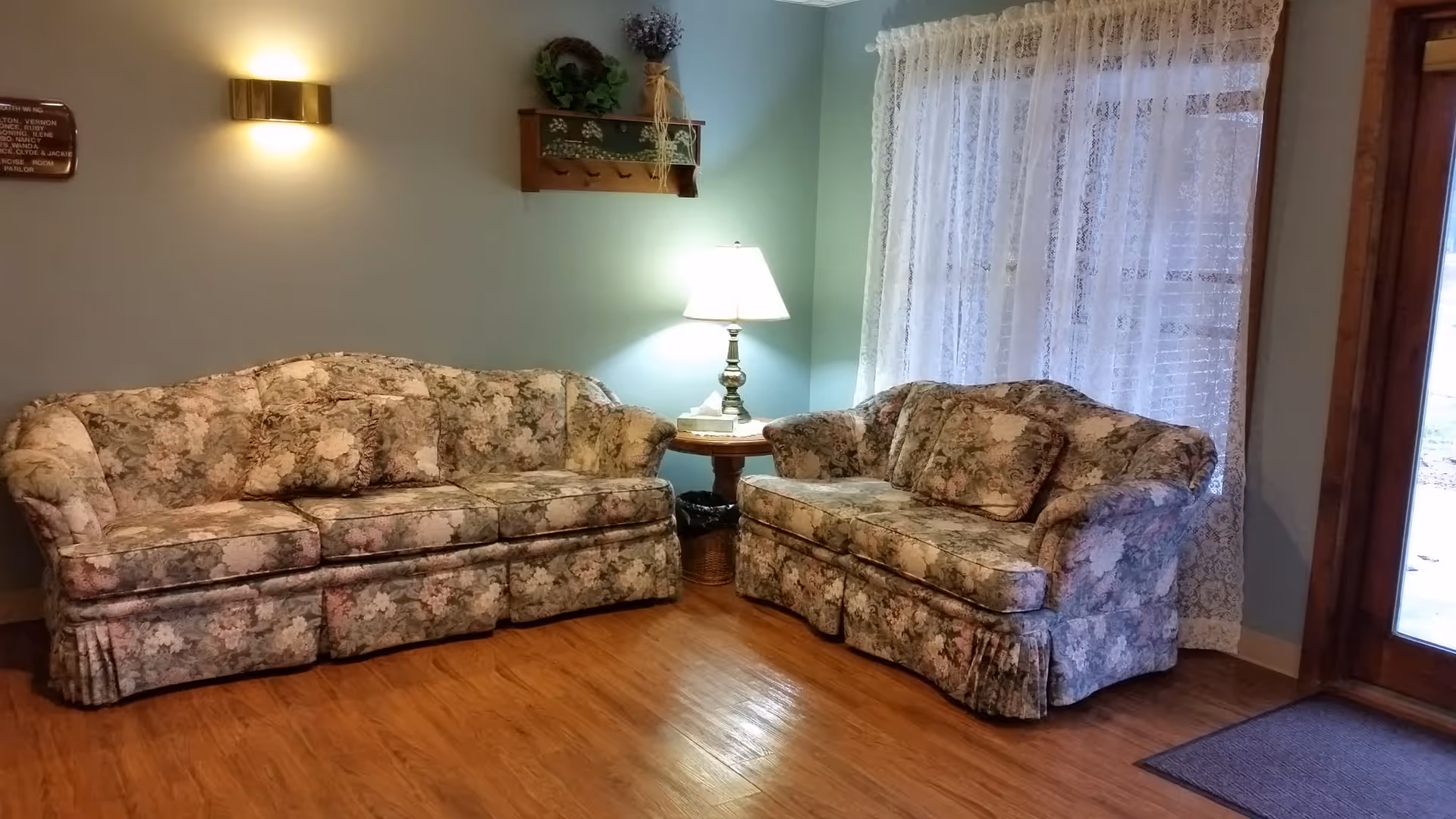 A cozy living room area with two floral-patterned sofas arranged around a wooden side table with a lamp. The walls are painted light blue, and there is a window with white lace curtains letting in natural light. The floor is wooden, and there is a door with glass panels on the right side.