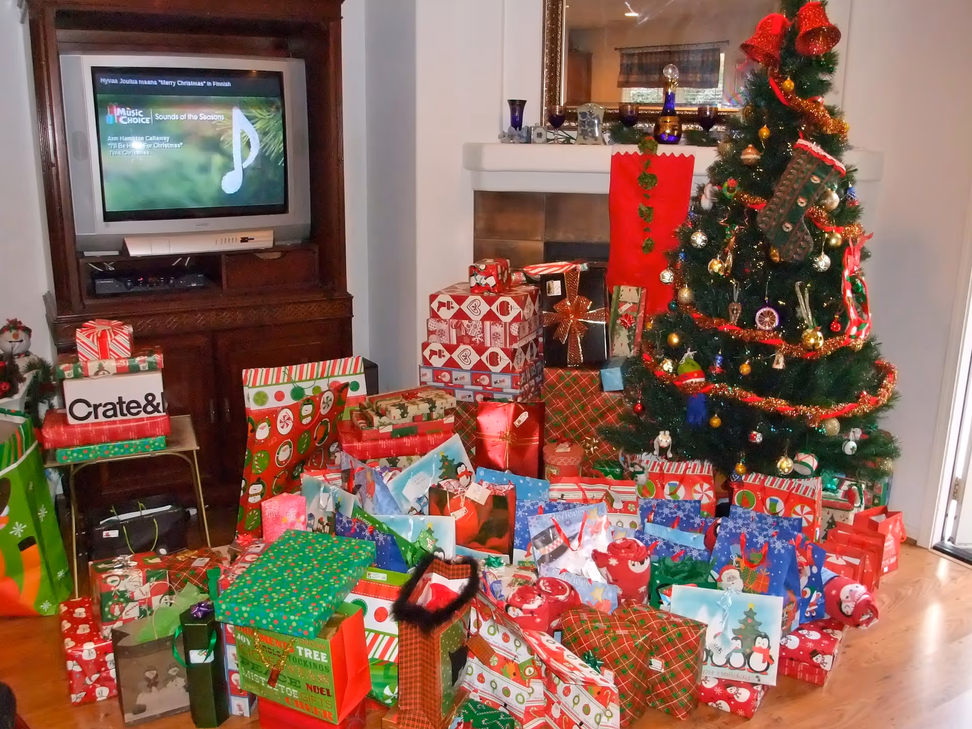 A living room decorated for Christmas with a Christmas tree adorned with ornaments, tinsel, and stockings. Numerous wrapped presents and gift bags are placed on the floor around the tree. A wooden entertainment center with a TV displaying a music channel is visible in the background.