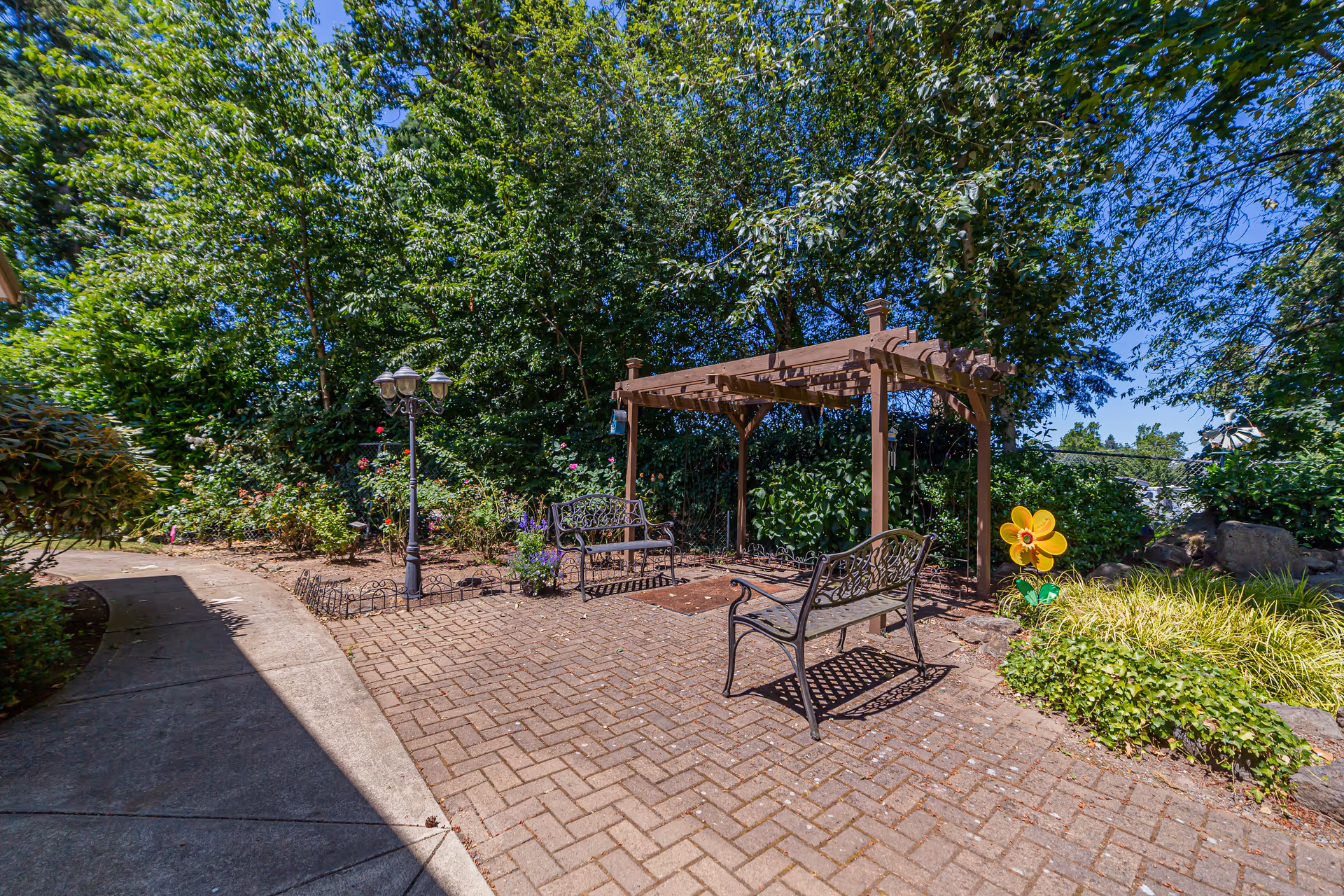 Outdoor garden area with a paved brick patio featuring two metal benches and a wooden pergola. Surrounding the patio are lush green trees, bushes, and flowering plants. A colorful decorative flower stake is visible in the garden bed to the right.