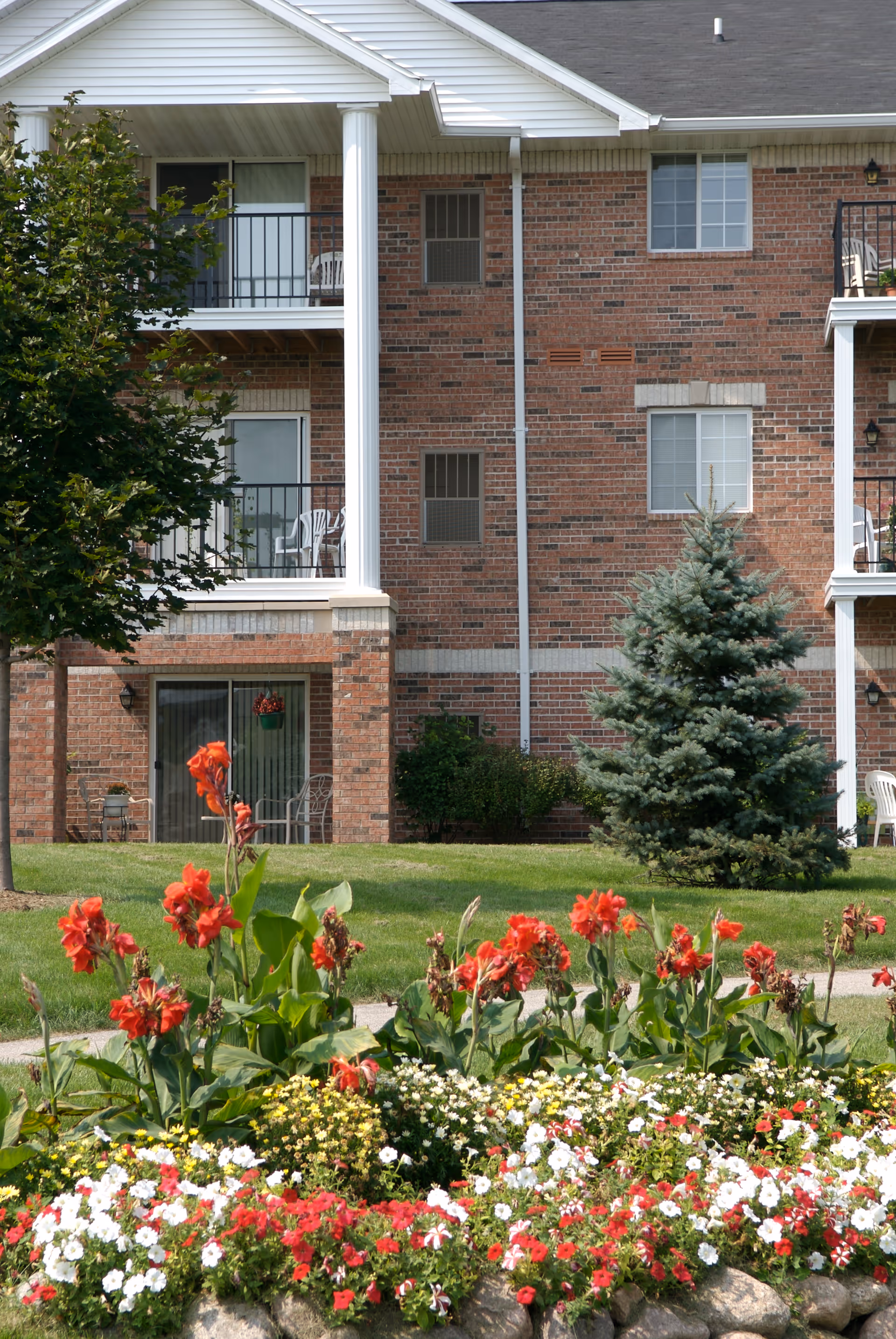 Brick apartment building facade with balconies, a pine tree, and colorful flowerbeds in the foreground.