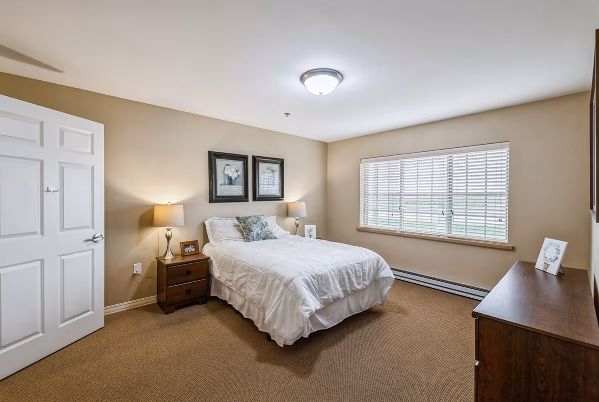 A neatly made bed with white bedding and decorative pillows in a bedroom with beige walls and carpet. There are two framed floral pictures above the bed, two bedside tables with lamps, a window with white blinds, and a wooden dresser with a small framed picture on it.