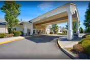 Entrance of a retirement community building with a covered drop-off area supported by white columns, surrounded by landscaped greenery and trees under a clear blue sky.
