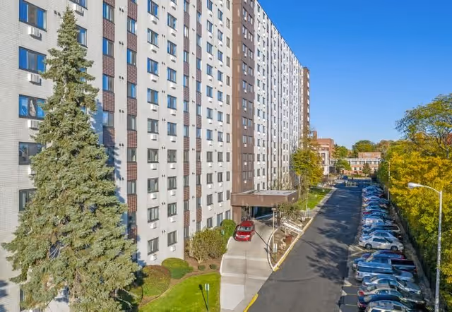 Exterior view of a tall multi-story senior living facility named River Towers with a driveway entrance, parked cars along the side, and trees with green and yellow foliage under a clear blue sky.