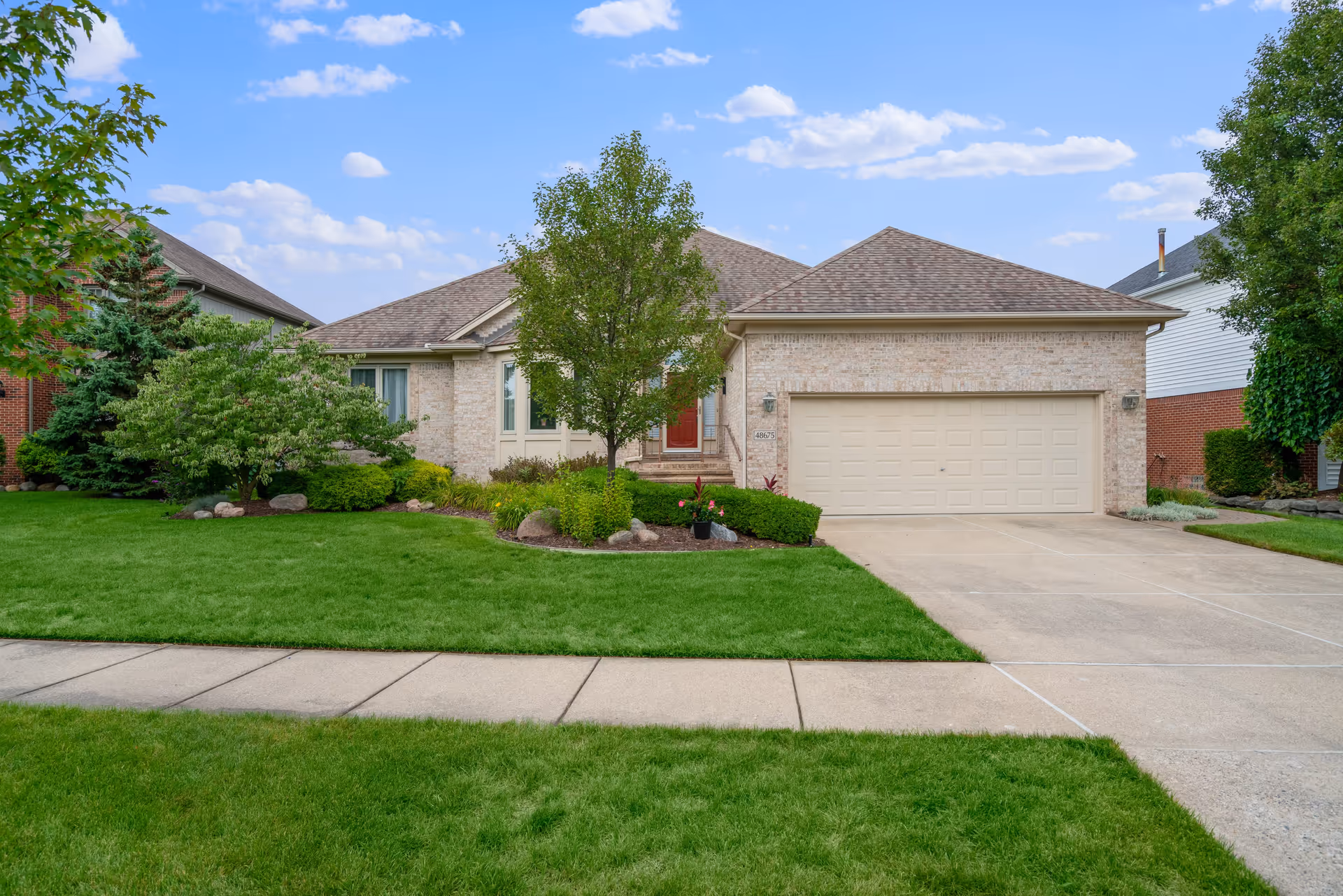Single-story brick house with a two-car garage, a red front door, and a well-maintained front yard with green grass, trees, and shrubs under a blue sky with scattered clouds.