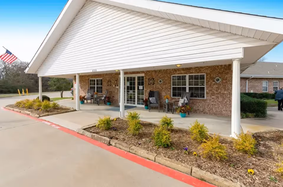 Front exterior view of a single-story brick building with a covered porch area featuring chairs and potted plants. There is a small landscaped area with shrubs and flowers in front of the porch, and an American flag is visible on the left side. A person is walking on the right side near the building.