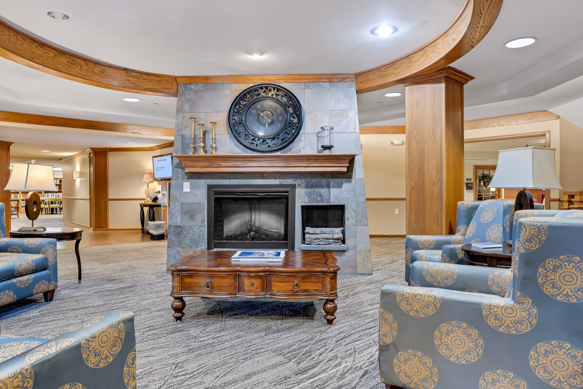 A cozy senior living facility common area with a stone fireplace as the focal point. The fireplace has a wooden mantel decorated with candlesticks and a large decorative clock above it. Surrounding the fireplace are patterned blue armchairs and wooden side tables with lamps. The room features wood trim and columns, carpeted flooring, and soft lighting, creating a warm and inviting atmosphere.