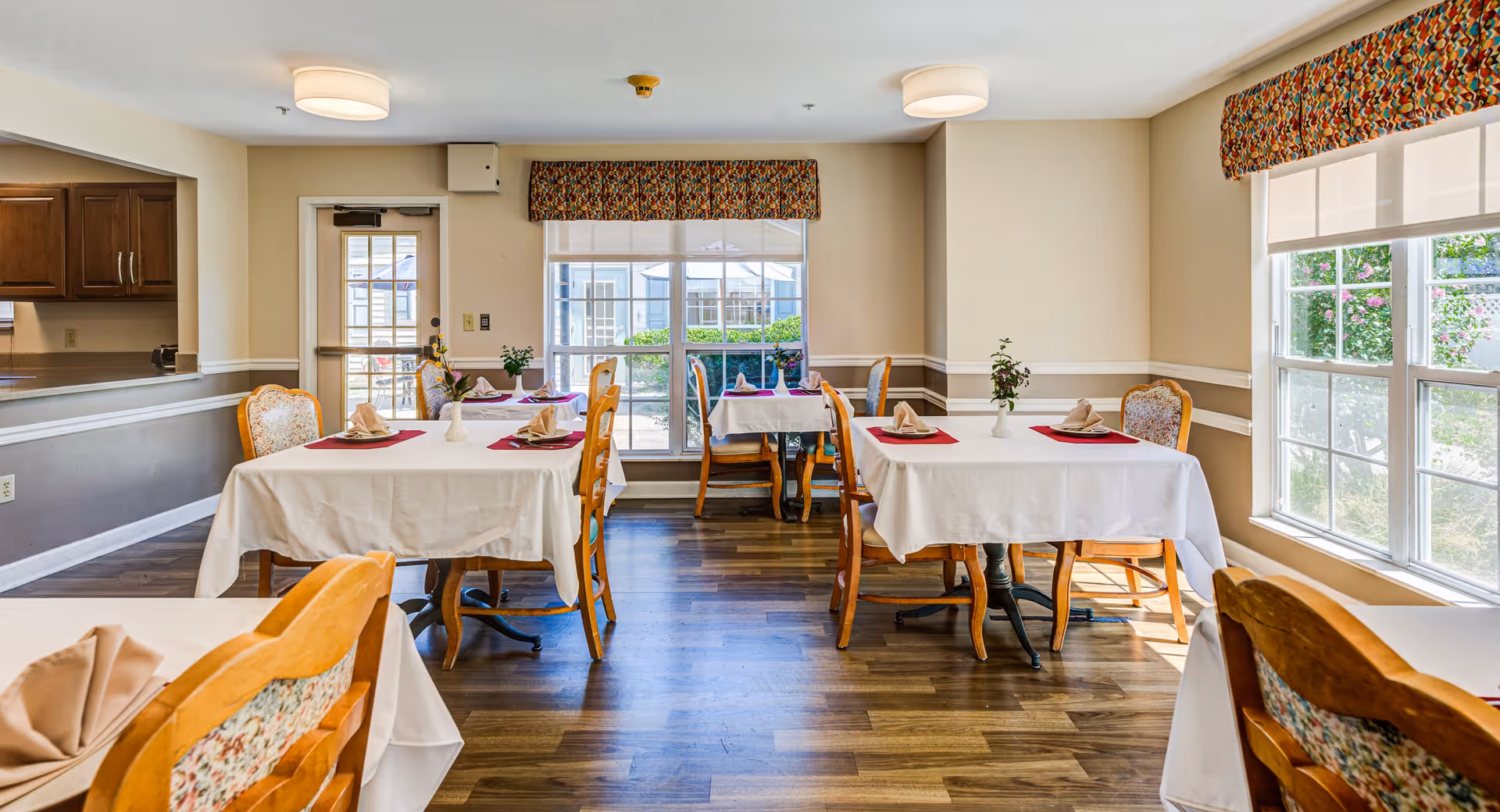 A bright dining room with several tables covered in white tablecloths and set with folded napkins and small vases with flowers. The room has large windows allowing natural light to fill the space, wooden chairs with patterned upholstery, and a door leading outside. The floor is wooden, and there is a kitchen pass-through window on the left side.