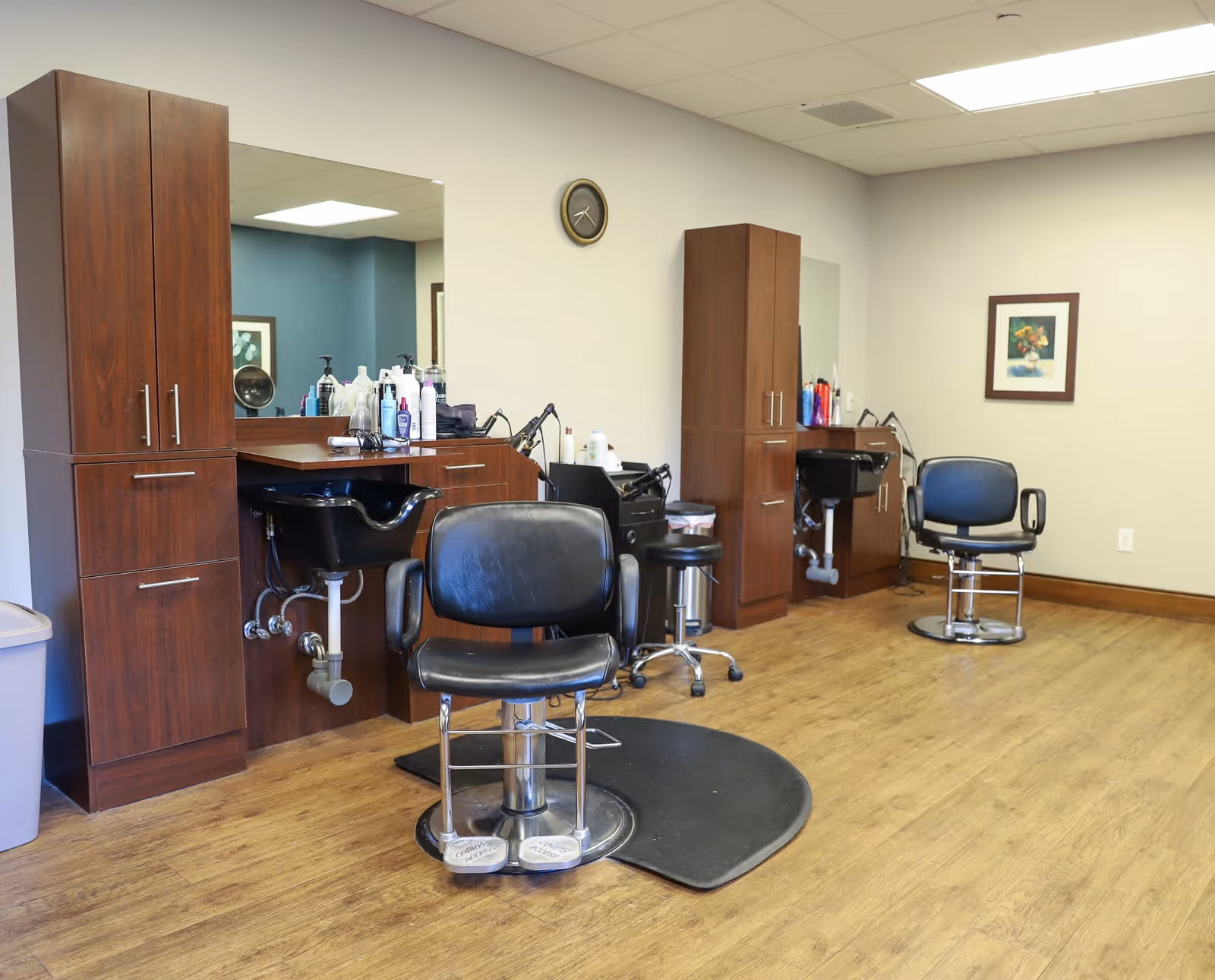 Interior of a hair salon area with two black salon chairs in front of wooden cabinets and sinks for washing hair. The room has wood flooring, a large mirror, a clock on the wall, and a framed picture.