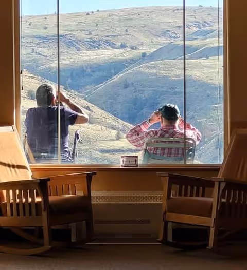 Two elderly men sitting outside on chairs looking through binoculars at a hilly landscape, viewed from inside a room with two wooden rocking chairs near a large window.