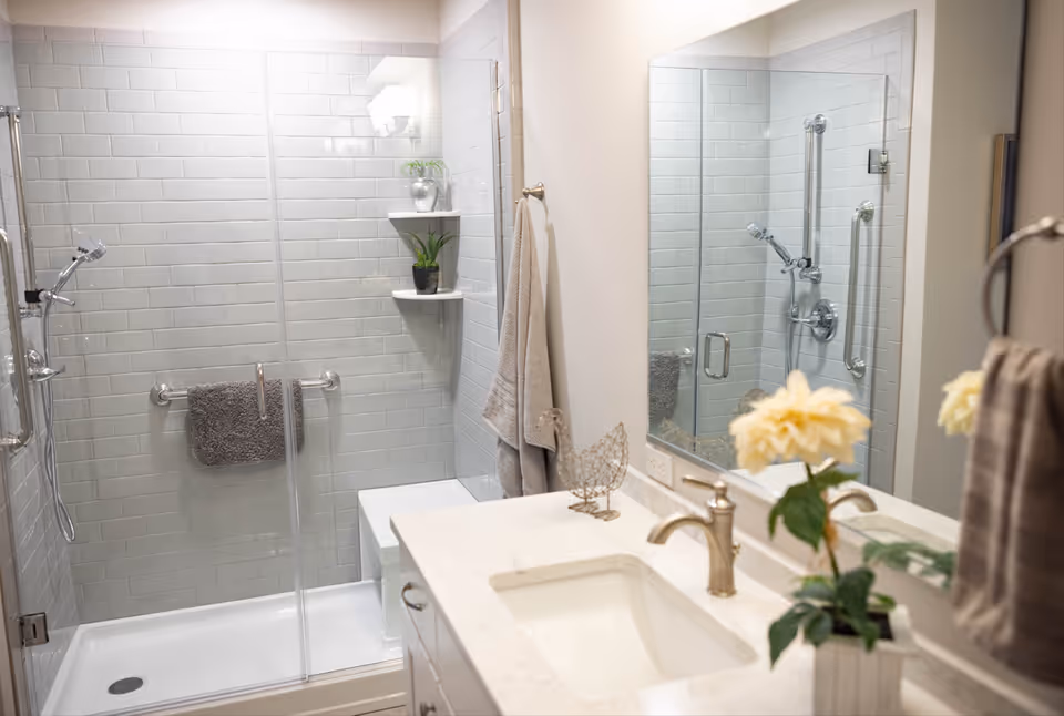 A clean and modern bathroom featuring a glass-enclosed shower with white subway tiles, a gray towel hanging on a grab bar inside the shower, and two small potted plants on corner shelves. The bathroom also has a white countertop with a built-in sink, a silver faucet, a decorative wire chicken, a yellow flower in a small vase, and a large mirror above the sink reflecting the shower area.