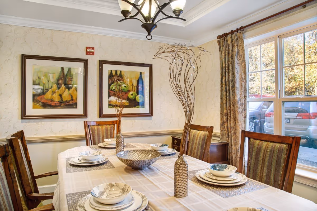 A dining room with a rectangular table covered with a beige tablecloth set with plates, bowls, and placemats. There are six wooden chairs with striped cushions around the table. Two decorative vases with tall, thin branches and a large decorative bowl are on the table. The room has patterned wallpaper, two framed paintings on the wall, a chandelier overhead, and a large window with floral curtains showing parked cars outside.
