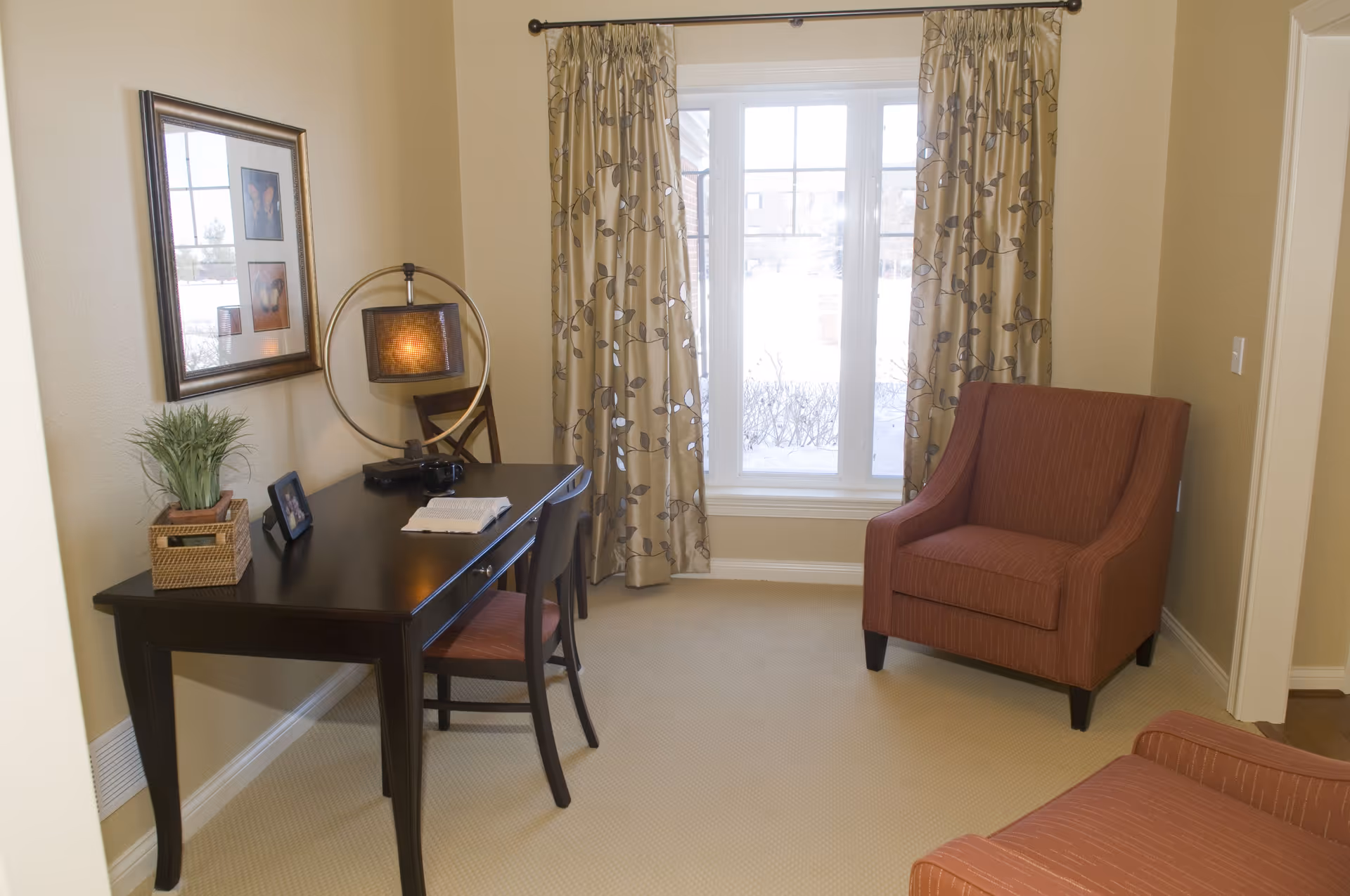 Sunlit sitting area with a desk, lamp, framed art, and an upholstered armchair by a window.