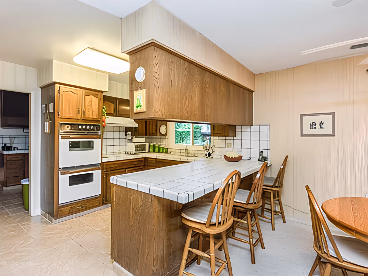 A kitchen area with wooden cabinets, white tiled countertops, and a breakfast bar with three wooden chairs. There is a double oven built into the cabinetry, a microwave on the counter, and a window above the sink showing greenery outside. Adjacent to the kitchen is a dining area with a round wooden table and chairs, and a framed picture on the wall.