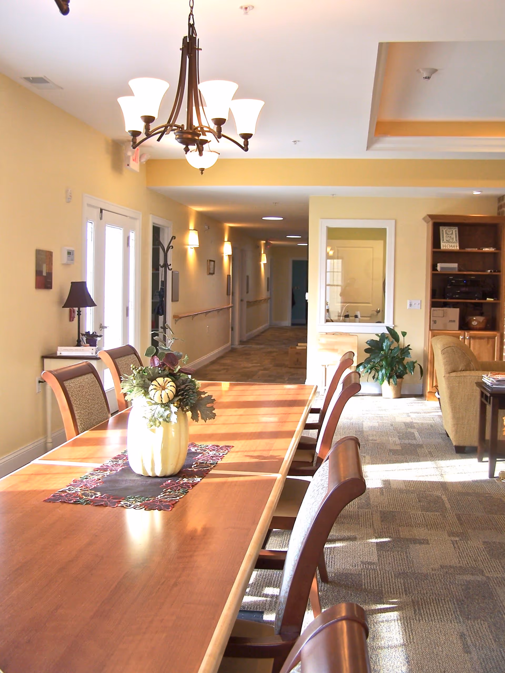 A bright and spacious dining area with a long wooden table surrounded by chairs. A decorative centerpiece with flowers and small pumpkins sits on the table. The room has beige walls, carpeted floors, and a chandelier hanging from the ceiling. In the background, there is a hallway with wall-mounted lights and a cozy seating area with a couch and bookshelf.