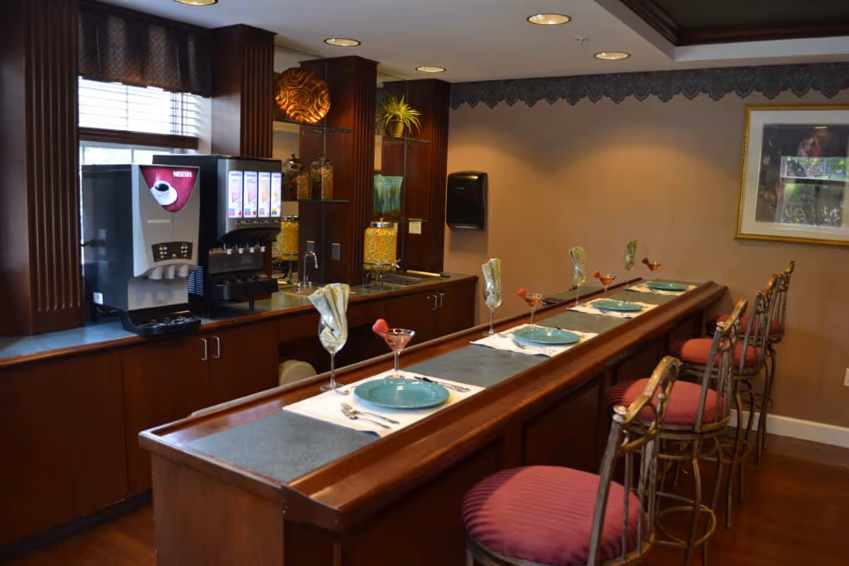 Long wooden bar counter set with plates, glasses, and bar stools in a small dining/kitchenette area with a beverage machine and cabinets.