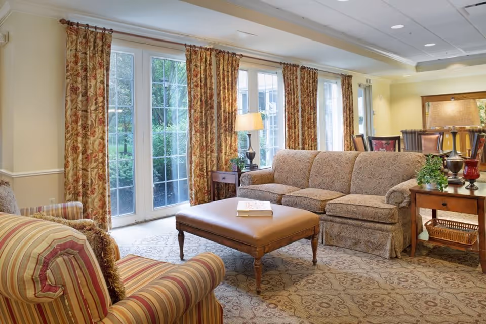 A cozy living room with a patterned beige sofa, a striped armchair, and a large ottoman in the center. The room features large windows with floral curtains allowing natural light to brighten the space. There are two side tables with lamps and a decorative plant, and a dining area with chairs is visible in the background.