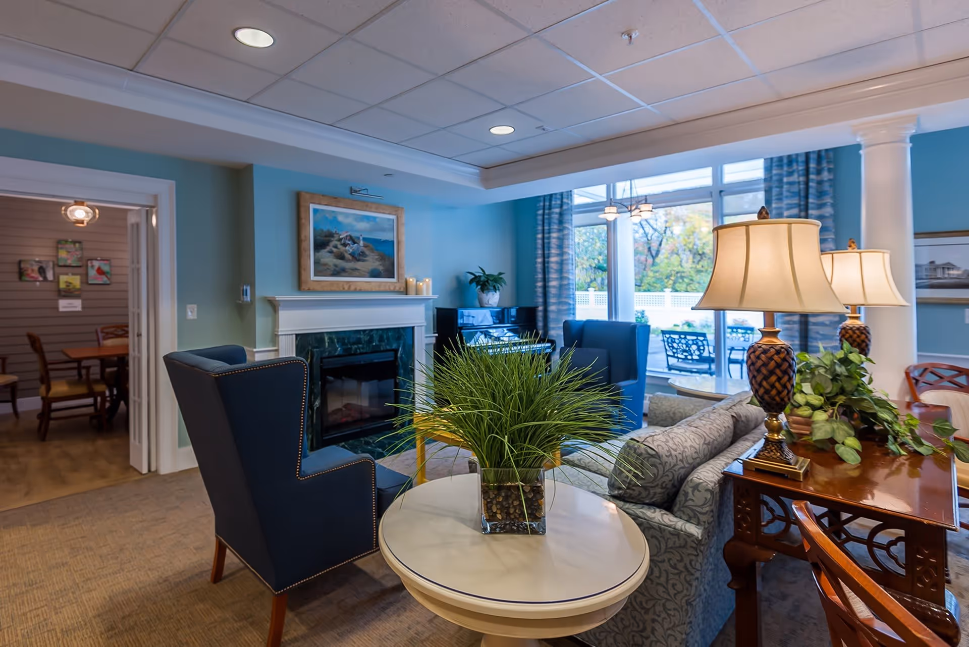 A cozy living room area with blue armchairs, a patterned sofa, a round white table with a green plant centerpiece, two table lamps with woven bases, and a fireplace with a painting above it. Large windows with blue curtains provide a view of an outdoor patio with chairs and a white fence.