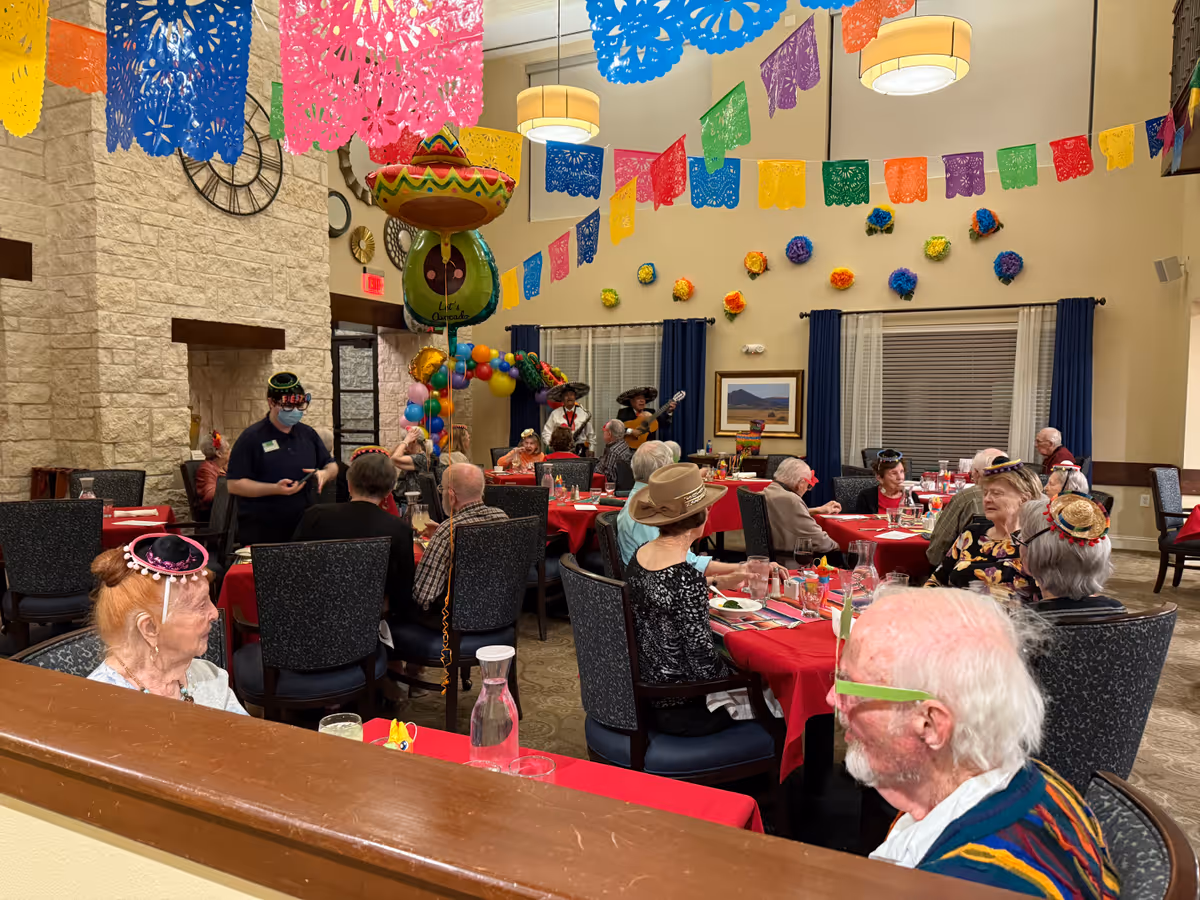 A festive dining room decorated with colorful paper banners and balloons. Elderly residents wearing small hats are seated at tables covered with red tablecloths, enjoying a meal and socializing. Two musicians in traditional attire are playing guitars in the background. The room has stone walls, large windows with blue curtains, and various wall decorations.