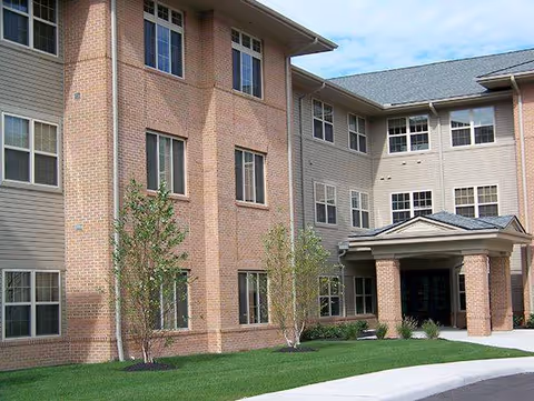 Brick senior living facility exterior with entrance, manicured lawn, and small trees.