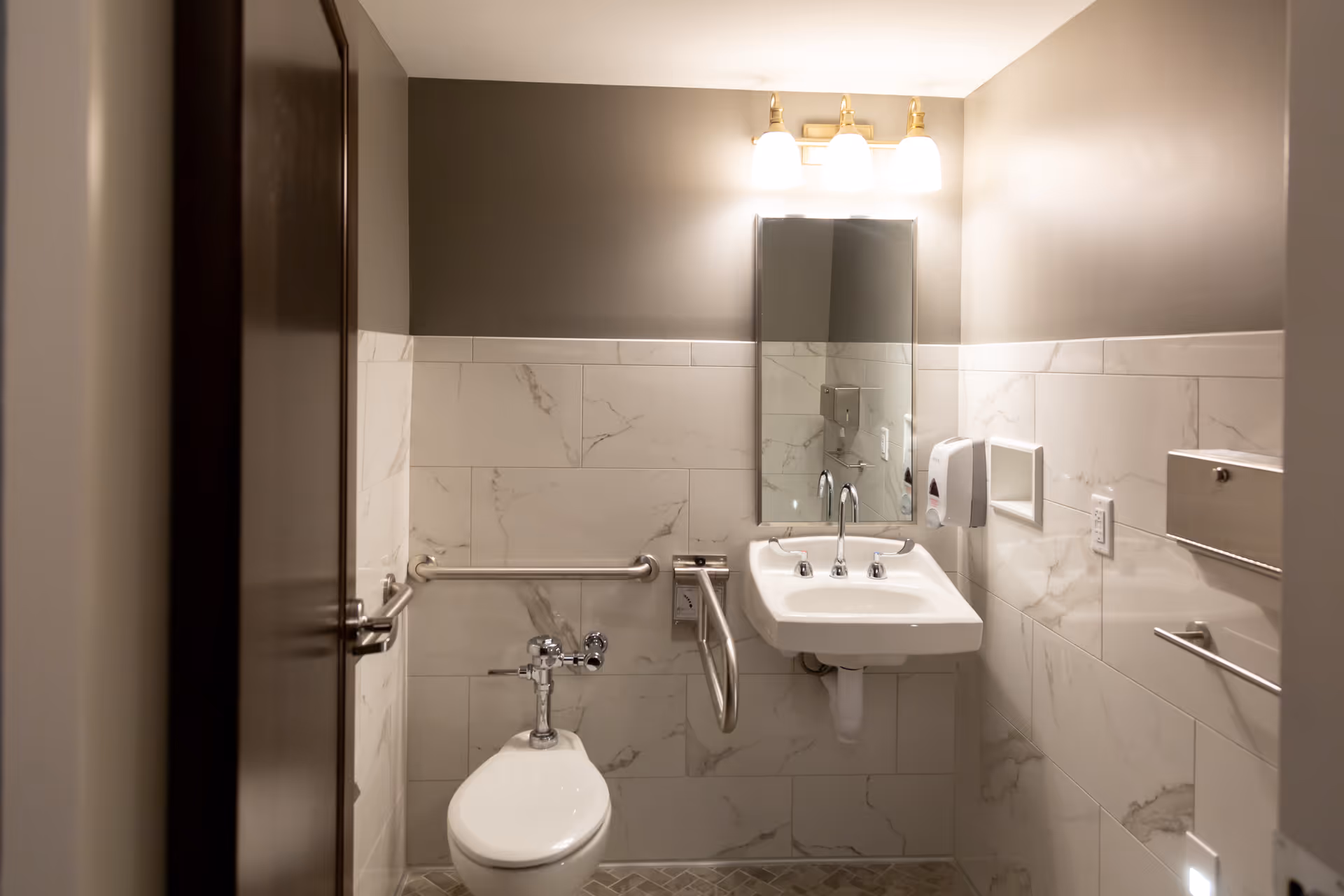 A small, clean bathroom with marble-patterned tiles on the walls and a gray painted upper wall. The bathroom features a white toilet with a metal grab bar on the left side, a white wall-mounted sink with a mirror above it, and a three-light fixture above the mirror. There is a soap dispenser mounted on the wall to the right of the sink and a metal paper towel dispenser further to the right.