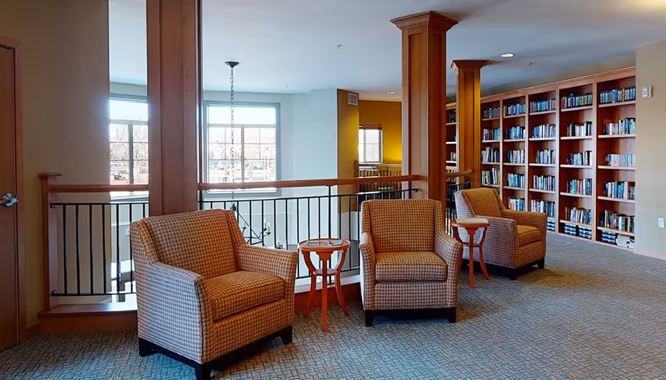 A cozy interior common area with three cushioned armchairs arranged in a row, each paired with a small wooden side table. Behind the chairs is a railing overlooking a lower level with large windows letting in natural light. To the right, there is a large built-in wooden bookshelf filled with books. The space has warm lighting and a carpeted floor.