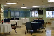 A spacious room set up with round tables covered in white and dark blue tablecloths, each surrounded by chairs. The room has large windows with green valances, a wall-mounted television, and ceiling lights. Decorations hang from the ceiling and walls, giving a welcoming atmosphere.