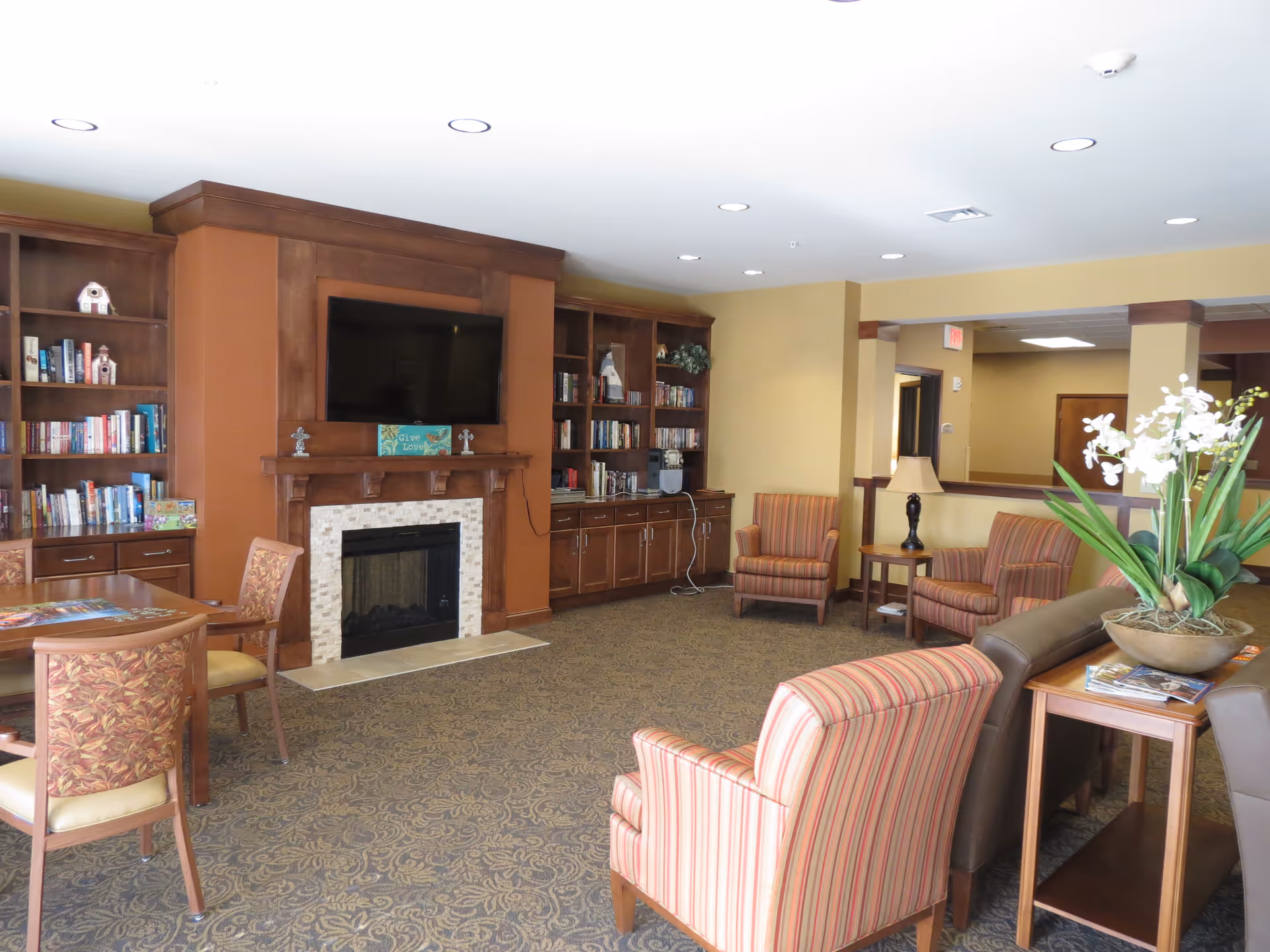 A cozy living room area in a senior living facility featuring a fireplace with a mounted flat-screen TV above it, built-in wooden bookshelves filled with books on either side, several upholstered armchairs with striped and floral patterns, a wooden table with chairs, a side table with a lamp, and a large plant with white flowers on a table in the foreground. The room has recessed ceiling lights and a patterned carpet.