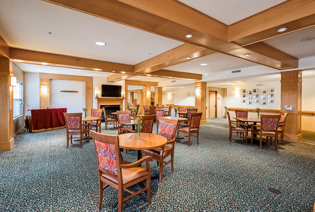 Bright communal dining/lounge area with round tables, patterned chairs, wood-beamed ceiling, and a fireplace with TV.